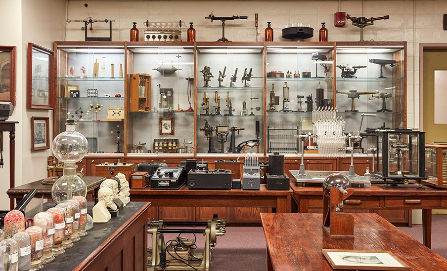The Oesper Apparatus Museum is shown with several lit wooden display cases filled with chemical apparatus, with additional artifacts displayed on top of the cases. One of two doors to the hall is propped open and several wooden tables also have artifacts on display.